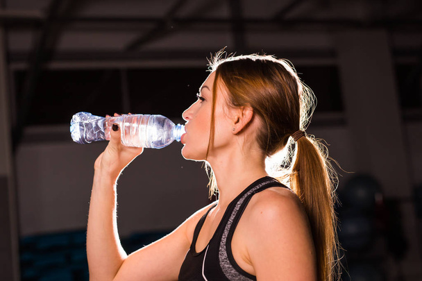 Фитнес молодая женщина пьет воду из бутылки. Мускулистая молодая женщина в спортзале отдыхает от тренировок
. - Фото, изображение