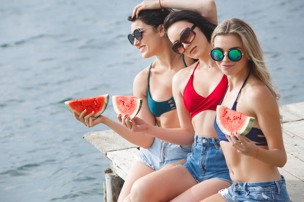 Group of young cheerful girls having fun on the pier eating watermelon and enjoying sun - Фото, изображение