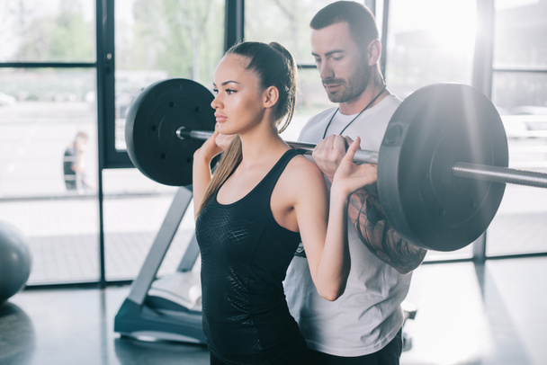male personal trainer helping sportswoman to do exercises with barbell at gym - Foto, Bild
