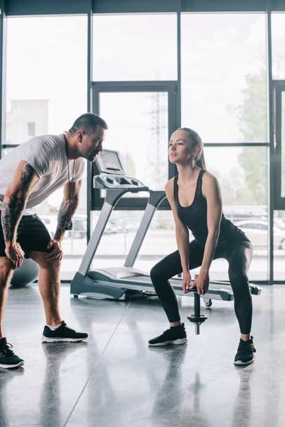 male personal trainer looking at sportswoman doing exercises with dumbbell at gym - Фото, зображення