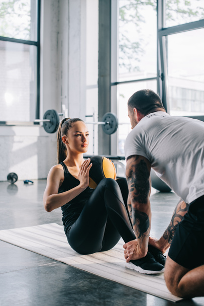 male personal trainer helping sportswoman to do abs with ball at gym - Fotó, kép
