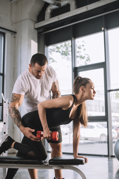 личный тренер мужского пола помогает спортсменке делать упражнения с гантелями в тренажерном зале
 - Фото, изображение