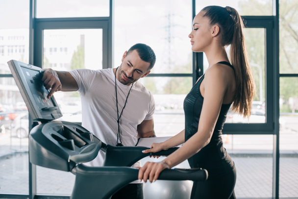 männlicher Personal Trainer zeigt auf Laufband-Bildschirm und Sportlerin steht in der Nähe im Fitnessstudio  - Foto, Bild