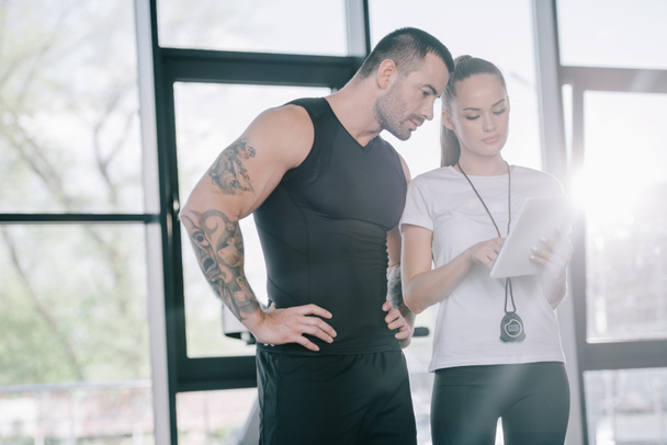 female personal trainer and sportsman looking at screen of digital tablet at gym - Photo, Image