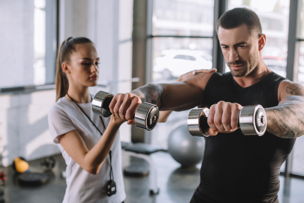 female personal trainer helping sportsman to do exercises with dumbbells at gym - Foto, afbeelding