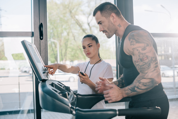 female personal trainer pointing at treadmill screen and running sportsman at gym - Photo, Image