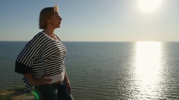 Dignified woman stands on the Black Sea shore and smiles in summer in slo-mo                              Profile of a charming blond woman in a waving blouse smiling happily and looking at the Black Sea at a splendid sunset in summer in slo-mo - Materiaali, video