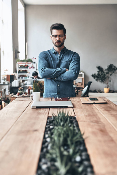 serious handsome young man looking at camera and keeping arms crossed while standing in creative working space, wooden table with plants and closed laptop     - Foto, Imagem