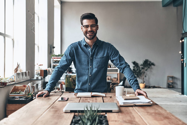 junger und erfolgreicher, gutaussehender junger Mann, der in die Kamera schaut und lächelt, während er im kreativen Arbeitsraum steht, geschlossener Laptop auf dem Tisch  - Foto, Bild
