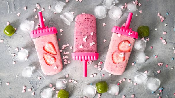 Summer heat: refreshing pink fruit ice cream with strawberry pieces surrounded by transparent and green ice cubes on a gray table. - Foto, afbeelding
