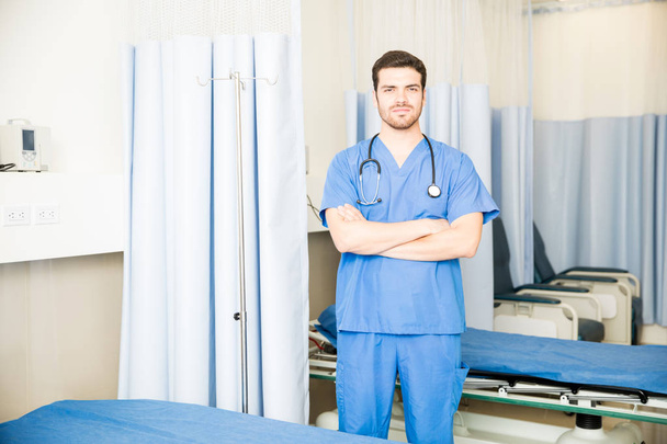 Aantrekkelijke jonge dokter in scrubs permanent met zijn armen gekruist in een ziekenhuis kamer met paar lege bedden - Foto, afbeelding