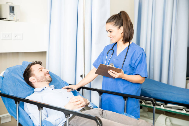 Uśmiechnięta lekarka ze schowka stojący obok na łóżku szpitalnym i rozmawia z pacjenta - Zdjęcie, obraz