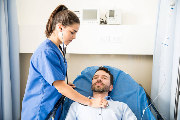 Enfermeira bonita usando um estetoscópio para examinar um paciente do sexo masculino em um quarto de hospital
 - Foto, Imagem