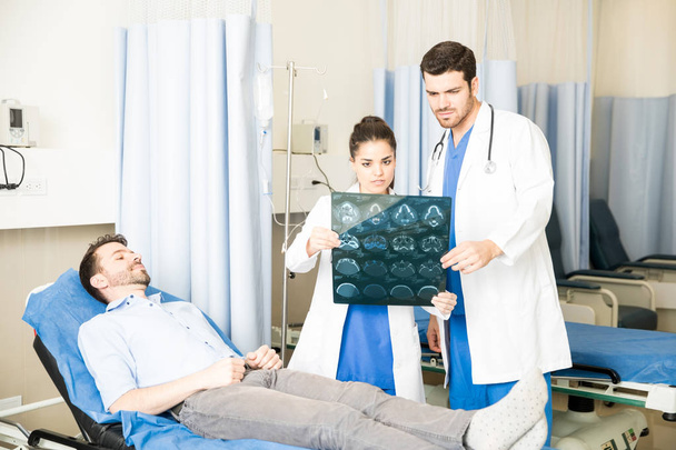 Два врача обсуждают результаты сканирования рентгеновского снимка с пациентом, лежащим на кровати в отделении скорой помощи
 - Фото, изображение
