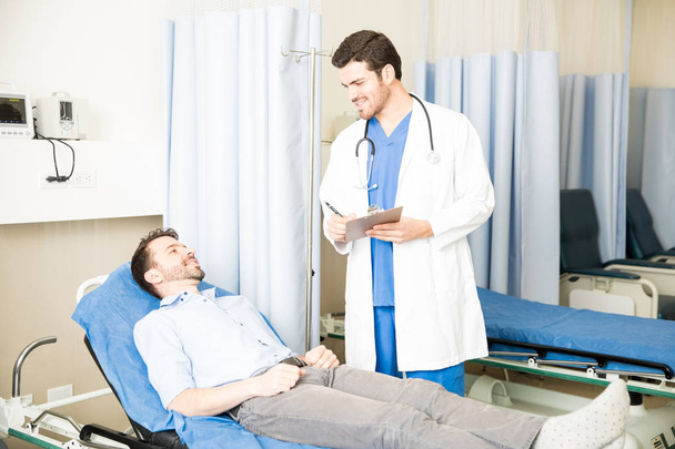Guapo doctor masculino con un portapapeles parado al lado de una cama y hablando con el paciente
 - Foto, Imagen