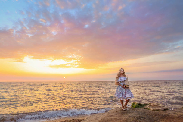 beautiful blonde girl holding wooden toy ship , standing on sea shore at sunset - Photo, Image