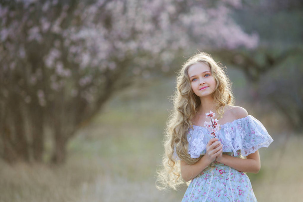 young beautiful blonde girl posing in blossoming garden wearing blue dress  - Fotografie, Obrázek