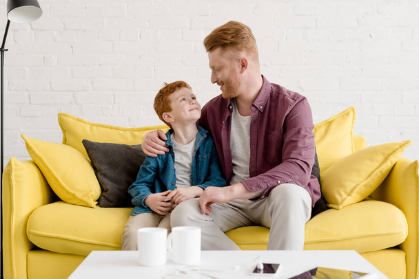 happy redhead father and son sitting together on sofa and smiling each other at home - Foto, Bild