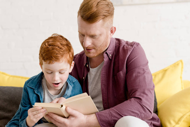 father looking at cute little son reading book at home - Photo, Image