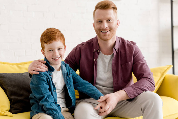 happy redhead father and son smiling at camera while sitting together on couch - Фото, зображення
