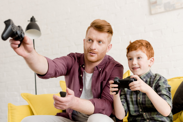 glimlachend roodharige vader en zoon op zoek weg tijdens het spelen met joysticks thuis - Foto, afbeelding