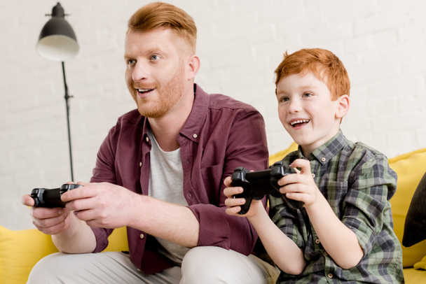 smiling father and son sitting on sofa and playing with joysticks at home  - 写真・画像