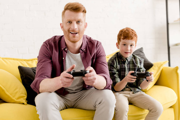 happy father and son sitting on sofa and playing with joysticks at home  - Photo, Image