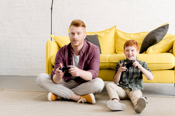 happy father and son sitting on carpet and playing with joysticks at home - Foto, afbeelding