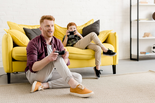 uśmiechający się czerwony haired ojca i syna, grając z joysticków razem w domu  - Zdjęcie, obraz
