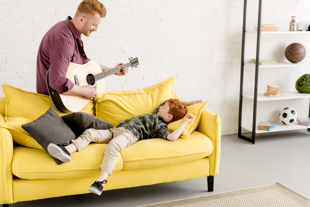 lachende vader spelen gitaar en schattig zoontje liggend op Bank  - Foto, afbeelding