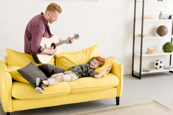 happy father playing guitar and cute smiling little son lying on couch  - 写真・画像