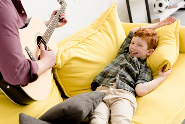 περικοπεί shot του πατέρα που παίζει κιθάρα, ενώ χαριτωμένο ευτυχείς γιος ξαπλωμένη στον καναπέ - Φωτογραφία, εικόνα