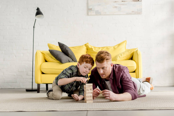 Konzentriert liegen Vater und Sohn auf Teppich und spielen zu Hause mit Holzklötzen - Foto, Bild