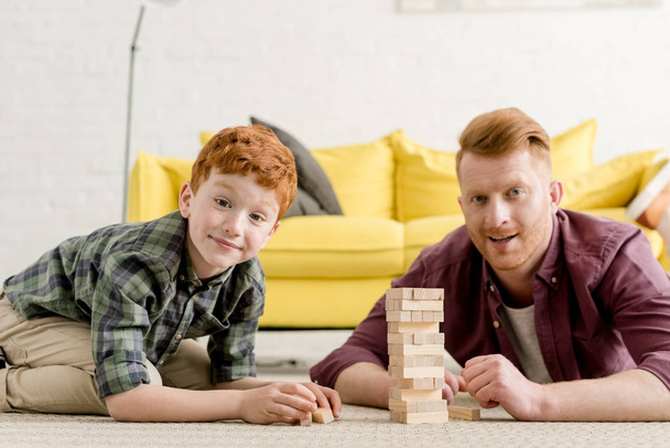 happy red haired father and son smiling at camera while playing with wooden blocks at home  - Zdjęcie, obraz