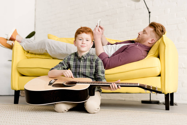 Netter kleiner Junge mit Akustikgitarrenwerbung, der in die Kamera lächelt, während der Vater das digitale Tablet auf dem Sofa hinter sich hat - Foto, Bild