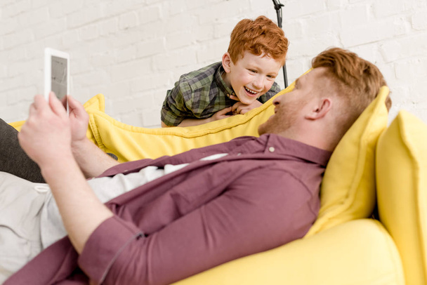 happy redhead man using digital tablet and looking at cute little son - Photo, image