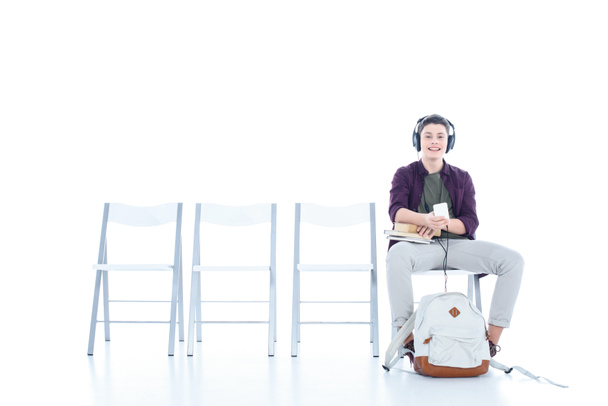 mosolyogva tizenéves diák fiú hallgat zenét, miközben ül a székre, elszigetelt fehér - Fotó, kép
