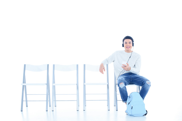 happy teenage student boy listening music while sitting on chair isolated on white - Photo, Image