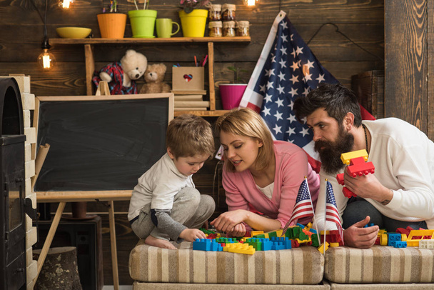 コンス トラクターを自宅で遊んで幸せなアメリカの家族。母と父のレンガで建設の構築を支援します。背景にアメリカ、アメリカ国旗とプレイルーム. - 写真・画像