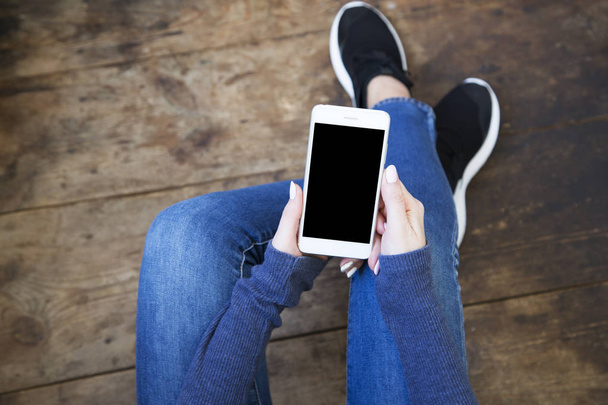La jeune fille est assise sur un plancher de bois et tient un smartphone avec un écran vide dans ses mains. Vue du dessus
. - Photo, image