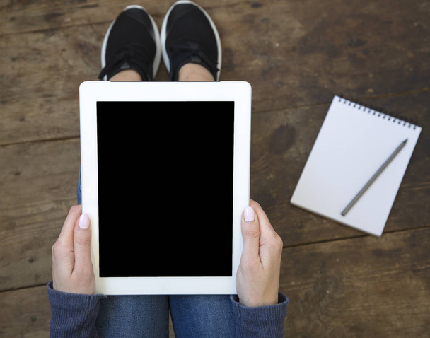 Het meisje zit op een houten vloer en een tablet met een leeg scherm houdt in haar handen. Laptop op de verdieping. Bovenaanzicht. - Foto, afbeelding