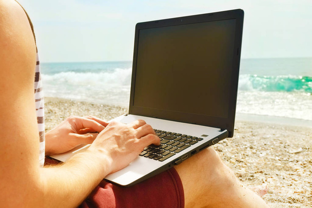 Young man in tank top, shorts, snapback cap coding on laptop at beach. Freelancer, hipster blogger, copywriter enjoying sea view. - Photo, Image