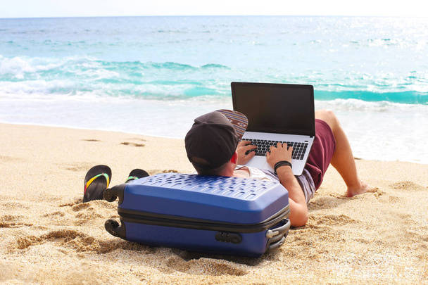 Young man in tank top, shorts, snapback cap at beach with laptop & suitcase. Freelancer, hipster blogger, writer enjoying sea view - Photo, Image
