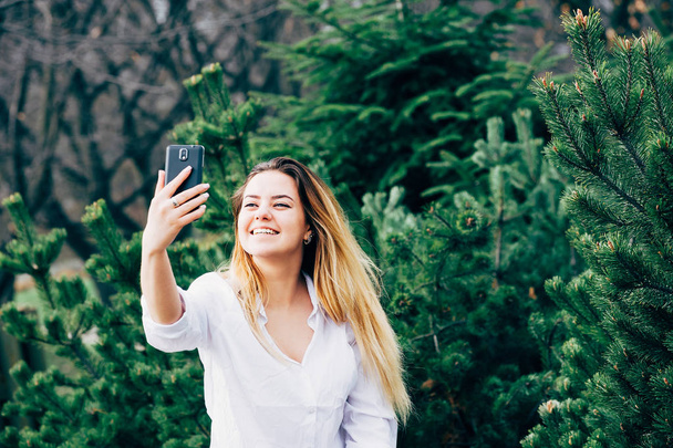 egy csinos, fiatal nő, mosolygott, és így selfie egy parkban - Fotó, kép