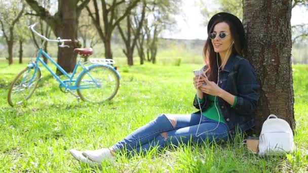 Cheerful and happy beautiful hipster woman in a hat and sunglasses listens to music with a phone in a city park and have fun on a retro bike background - Filmati, video