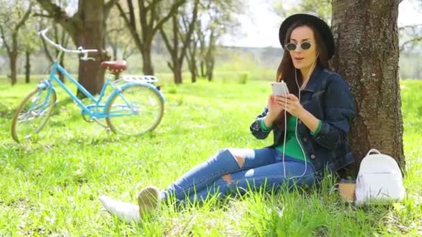 Cheerful and happy beautiful hipster woman in a hat and sunglasses listens to music with a phone in a city park and have fun on a retro bike background - Filmati, video