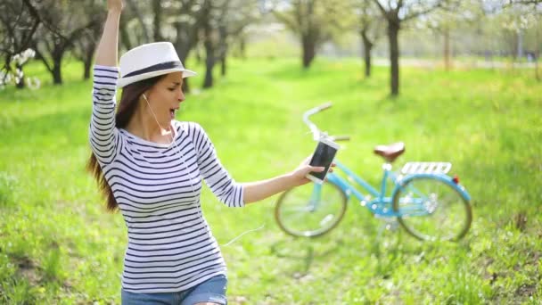 Cheerful and happy beautiful young woman in a hat and casual wear listens to music with a phone in a city park and dances on a retro bike background - Materiaali, video