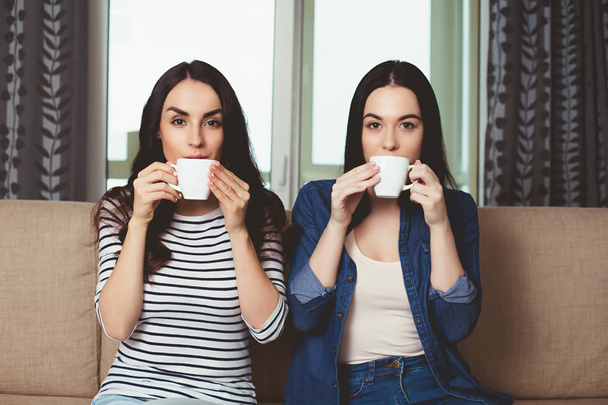 Two young beautiful smiling girls in casual clothes communicate and drink a coffee or tea sitting on the sofa at home. - Zdjęcie, obraz