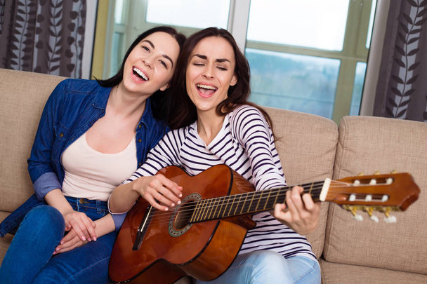 Cheerful beautiful girl friends playing guitar sitting on the couch. - Fotó, kép