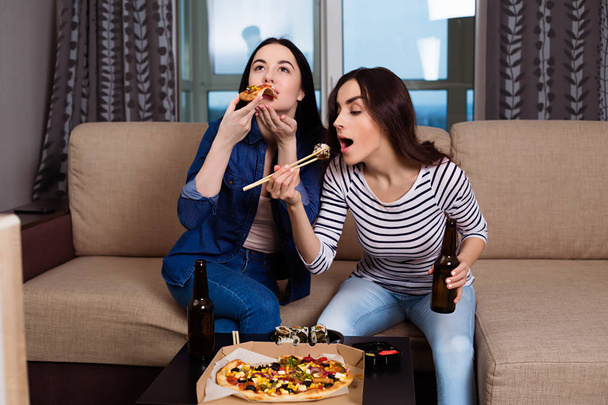 Relax on the couch with friends. Two beautiful young smiling girlfriends at home on the couch watching movies and eating rolls and pizza. - Φωτογραφία, εικόνα